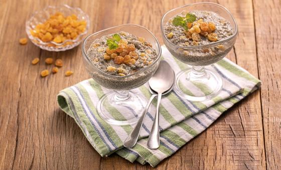 Foto da receita de Pudim de Chia com Uva Passa. Observa-se duas taças de vidro transparentes com o pudim e as uvas, sobre um guardanapo listrado em tons de verde claro. Também há duas colheres sobre o guardanapo e, atrás, há um pote com uvas passas.
