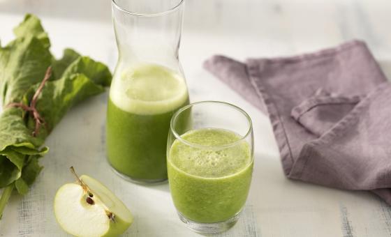 Fotografia em tons de verde e roxo de uma bancada branca, sobre ela dois copos de vidro com suco verde. Ao lado um paninho roxo, uma fatia de maçã verde e folhas de couve.