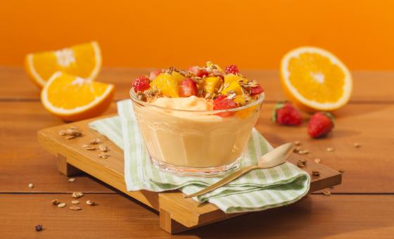Fotografia em tons de laranja em uma bancada de madeira, uma tábua de madeira, um paninho verde, um recipiente de vidro com o iogurte de laranja, morango e granola. Ao fundo, laranja e morango.