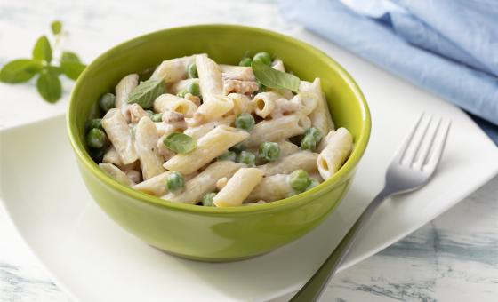 Fotografia em tons de branco, verde e azul de uma bancada branca e um prato retangular branco, sobre ele um recipiente redondo verde com o macarrão e um garfo. Ao fundo um paninho azul e folhas de manjericão.