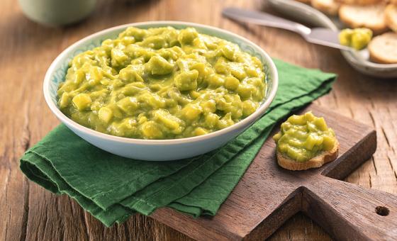 Foto da receita de Guacamole. Observa-se um pote redondo com a guacamole, sobre um guardanapo verde e uma tábua de madeira. Ao lado do recipiente, uma torrada redonda com o guacamole. As mesmas torradas decoram o fundo da foto.