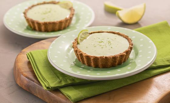 Foto da receita de tortinha de limão. Observa-se dois pratos verdes de sobremesa com as tortinhas sobre um guardanapo verde e uma tábua de madeira. Atrás, duas fatias de limão.