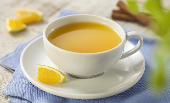Fotografia em tons de azul, cinza e amarelo de uma bancada cinza com um paninho azul e sobre ele um prato branco redondo com uma xícara com chá de laranja. Ao fundo paus de canela e pedaços de laranja.