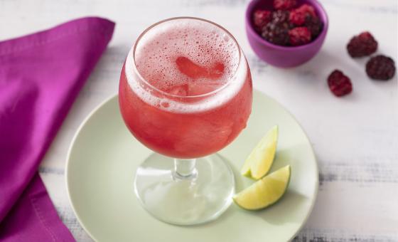 Fotografia em tons de branco, roxo e verde de uma bancada de madeira branca e um prato redondo verde, sobre ele uma taça com o refresco de tônica e duas fatias de limão. Ao lado um paninho roxo e um recipiente redondo roxo com frutas vermelhas.