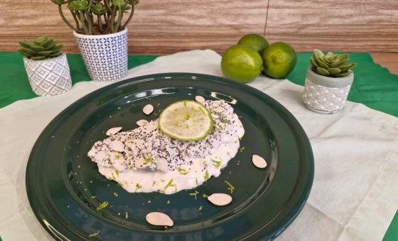 Foto em tons de branco da receita de tapioca servida com uma rodela de limão por cima, sobre um prato preto em cima de uma toalha de linho com limões e vasinhos de planta ao lado