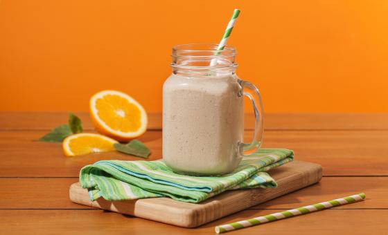 Fotografia em tons de laranja em uma bancada de madeira, um fundo laranja. Ao centro, uma tábua de madeira, um paninho verde, um canudo de papel verde e em cima um copo grande de vidro com o shake de banana dentro. Ao fundo, uma laranja aberta e folhas.