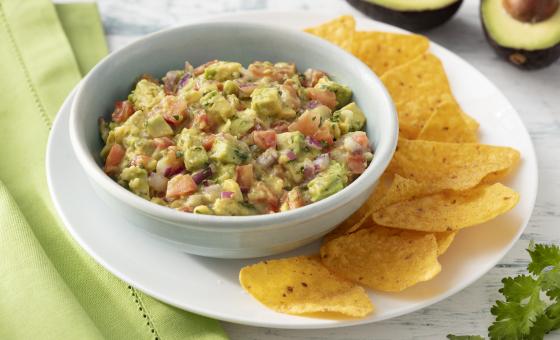 Fotografia em tons de branco e verde de uma bancada de madeira branca com um paninho verde e um prato redondo branco, sobre ele um recipiente branco redondo com a guacamole e nachos. Ao fundo um avocado e folhas de salsinha.