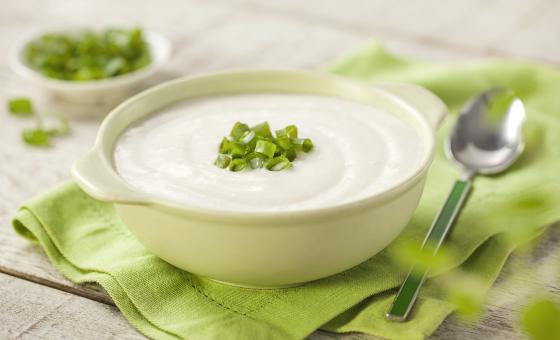 Fotografia em tons de verde em uma bancada de madeira com um guardanapo de pano verde ao centro com uma tigelinha branca e o creme de couve-flor dentro enfeitado com cebolinhas verdes. Ao lado, uma colher e ao fundo um potinho com cebolinhas verdes.