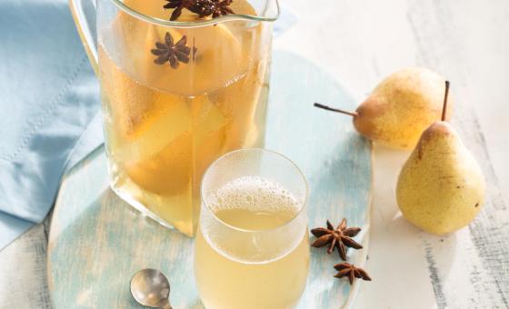 Fotografia em tons de azul em uma bancada de madeira cinza clara, um pano azul, uma bandeja de madeira azul mesclada com uma jarra de vidro e o refresco de pera e erva-doce dentro dele. Ao lado, um copo de vidro com a bebida, peras e anis-estrelado.