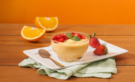 Fotografia em tons de laranja em uma bancada de madeira, um fundo laranja, um recipiente branco retangular, um pano verde claro e uma tacinha de vidro com a mousse de laranja dentro dela, com morangos para decorar. Ao fundo, uma laranja partida ao meio.