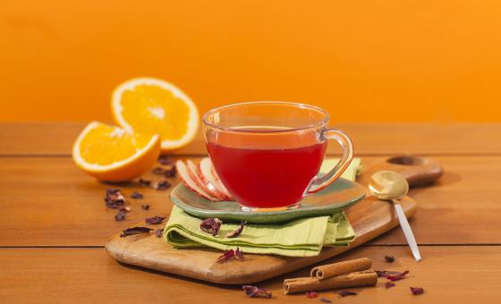 Fotografia em tons de laranja em uma bancada de madeira e um fundo laranja. Ao centro, uma tábua de madeira, um paninho verde e uma xícara de vidro com o chá de hibisco dentro dele. Ao fundo, laranja e canela em pau espalhados pela bancada.