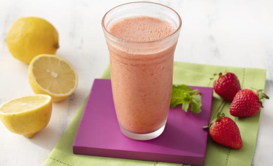 Fotografia em tons de branco, roxo e verde de uma bancada de madeira branca e um paninho verde, sore ele um apoio de copo quadrado roxo com um copo de vidro com o suco de morango. Ao lado três morangos e dois limões.