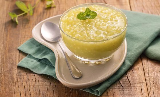 Foto da receita de Sagu Suco Verde. Observa-se uma taça transparente contendo o sagu sobre um prato e um guardanapo verde . Ao lado, uma colherinha e um ramo de hortelã.
